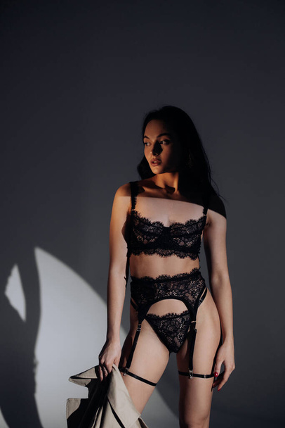 Jovem mulher sexy posa de pé em lingerie rendada preta no estilo BDSM com capa na mão contra fundo escuro. - Foto, Imagem
