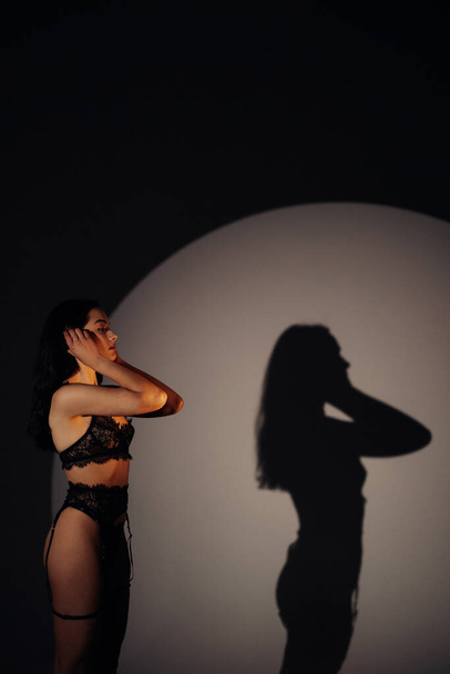 Young sexy woman poses in dark in black lacy lingerie in BDSM style against background of her shadow on the wall. She is illuminated. - Foto, afbeelding