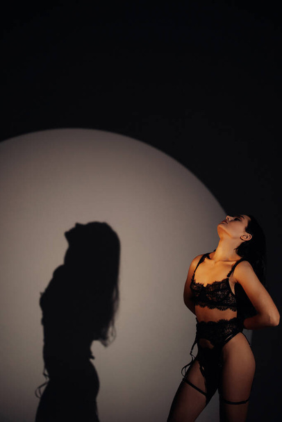 Молода сексуальна жінка позує в темній білизні чорного мережива в стилі БДСМ на тлі її тіні на стіні. Вона освітлена
. - Фото, зображення