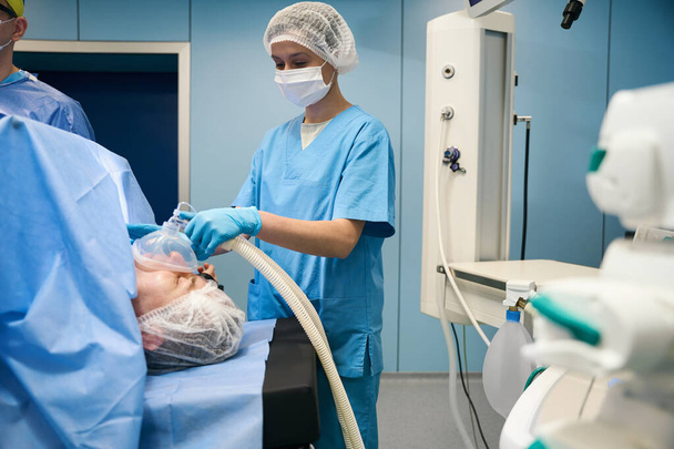 Женщина-анестезиолог держит маску с анестезией перед лицом пациента, ее коллеги работают рядом - Фото, изображение