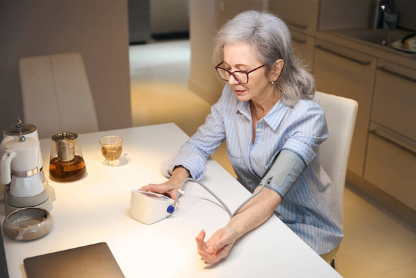 Lonely pensioner measures her blood pressure with a modern tonometer, she sits in the kitchen at the table - Photo, image