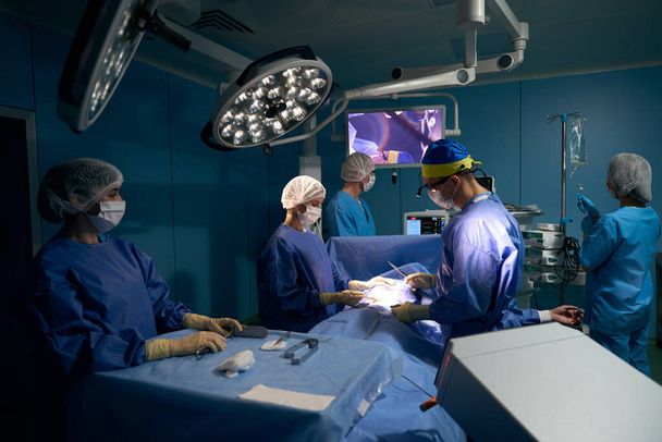Chirurg z aparatem na głowie operuje pacjenta na nowoczesnej sali operacyjnej, pacjent pod narkozą leży na stole - Zdjęcie, obraz