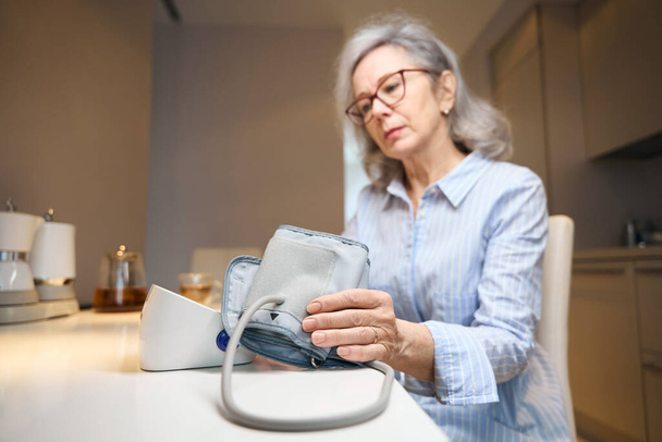 Elderly tired woman uses a tonometer to monitor blood pressure, she sits at home in the kitchen - Foto, Imagen