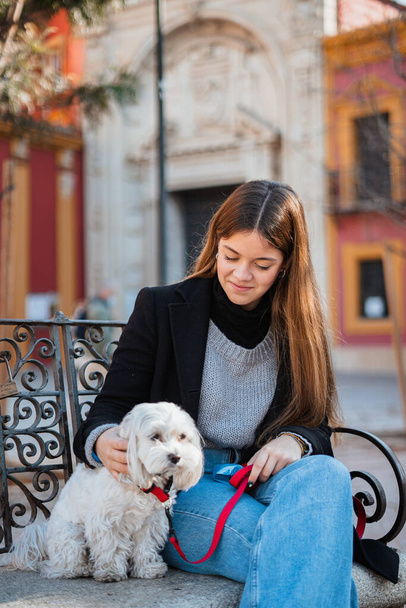 Χαμογελώντας κορίτσι κοιτάζοντας το σκύλο της, ενώ αυτοί κάθονται σε ένα παγκάκι στην πόλη - Φωτογραφία, εικόνα