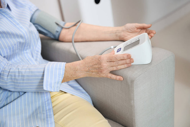 Woman sits on a couch and measures blood pressure with a tonometer, she is unwell - Photo, Image