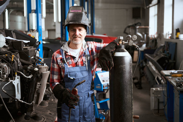 Employee of a car repair shop stands in a workshop with welding machine, a man leaned on a gas cylinder - 写真・画像