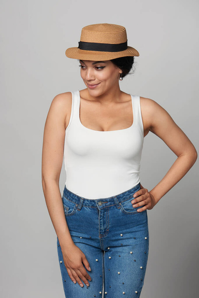 Relaxed casual mixed race african american - caucasian woman in jeans and white tank top wearing canotier straw hat, looking down smiling - Photo, image