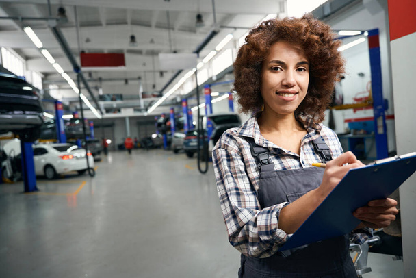 Woman auto mechanic in a repair shop fills out paperwork, she has a blue folder in her hands - Foto, imagen