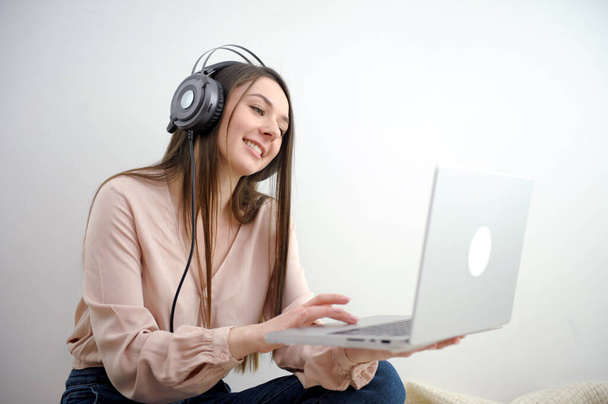 Vidám kaukázusi nő fülhallgató, amelynek online video hívás a laptop, ül a széken Millennial hölgy részt webinar, kommunikál távolról az interneten szép mosoly kamera lassan zoom - Fotó, kép
