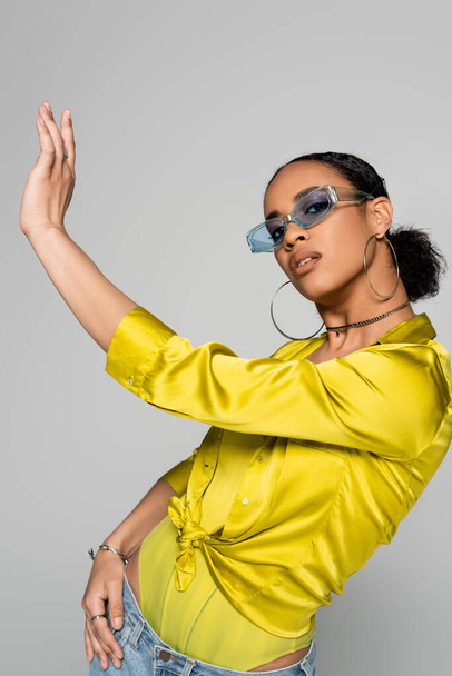 charming african american woman in blue sunglasses and stylish outfit posing with outstretched hand isolated on grey   - Foto, immagini