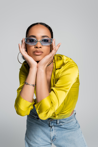 pretty african american woman in blue sunglasses touching face and looking at camera isolated on grey - Fotografie, Obrázek