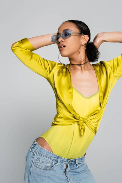 brunette african american model in blue sunglasses posing and looking away isolated on grey  - 写真・画像