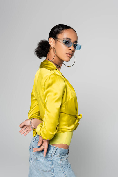 young african american model in blue sunglasses and trendy clothes posing with hands behind back isolated on grey  - Fotografie, Obrázek