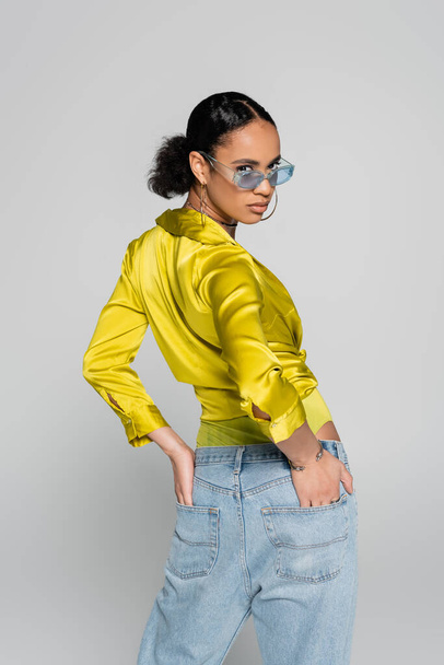 young african american model in blue sunglasses and trendy clothes posing with hands in back pockets isolated on grey  - Foto, afbeelding