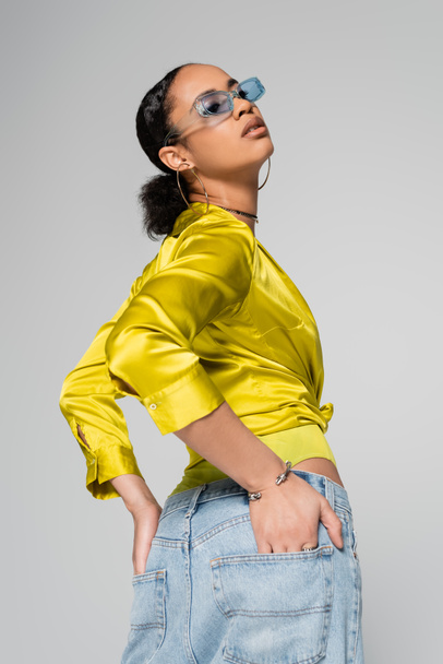 african american model in blue sunglasses and stylish clothes posing with hands in pockets isolated on grey  - Fotografie, Obrázek