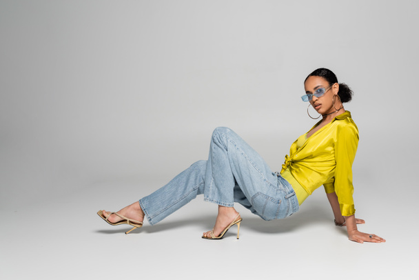 Yüksek topuklu ve gri arka planda poz veren modaya uygun gözlüklü Afro-Amerikan kadın.  - Fotoğraf, Görsel