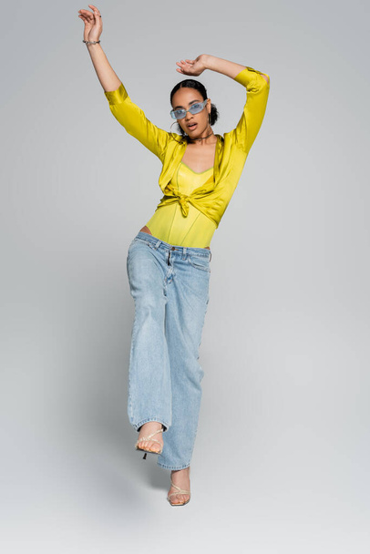 full length of surprised african american model in trendy clothes posing on one leg on grey  - Fotó, kép