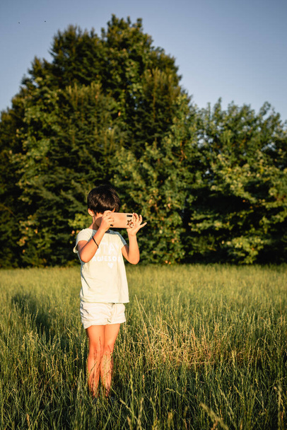 bambino in piedi con i capelli corti in campo verde sera scattare foto con il telefono cellulare - Foto, immagini