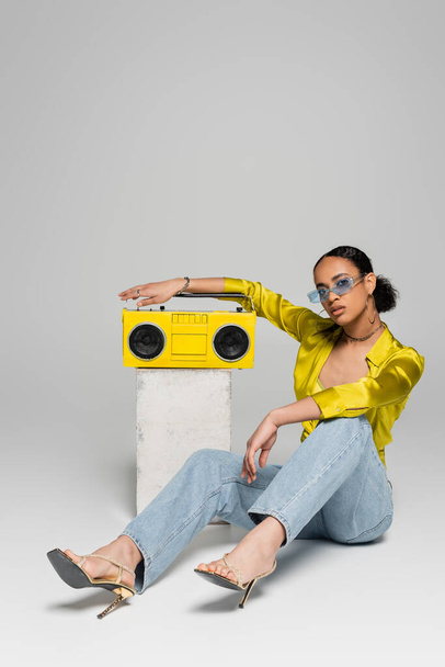 full length of brunette african american woman in sunglasses sitting near concrete cube and retro boombox on grey  - Foto, imagen
