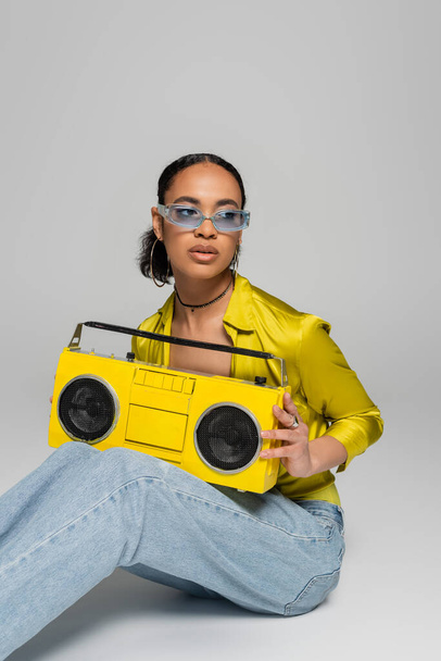 stylish african american woman in blue sunglasses sitting and holding retro boombox on grey  - Foto, immagini
