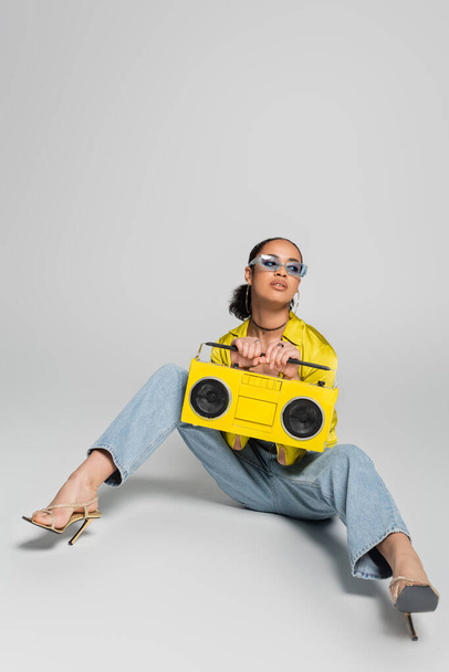 full length of brunette african american woman in blue sunglasses posing with yellow boombox on grey  - Foto, Bild