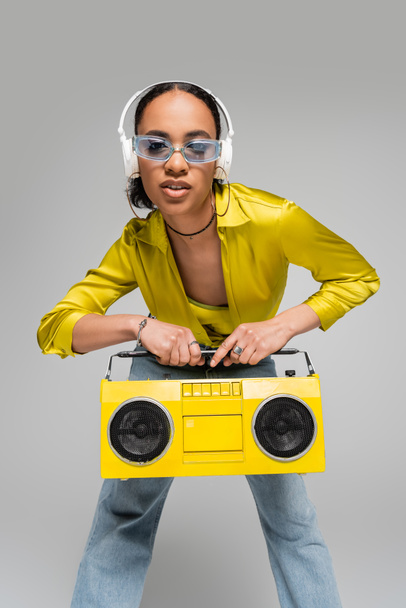 stylový africký americký žena poslech hudby v bezdrátových sluchátkách při držení boombox izolované na šedé  - Fotografie, Obrázek