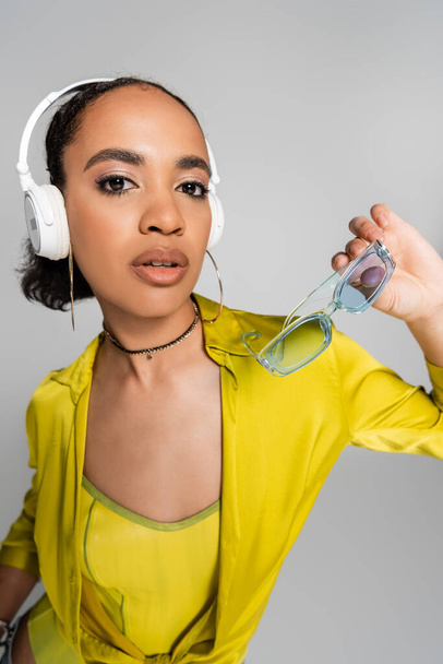 csinos afro-amerikai nő hallgat zenét vezeték nélküli fejhallgató és kezében kék napszemüveg elszigetelt szürke  - Fotó, kép