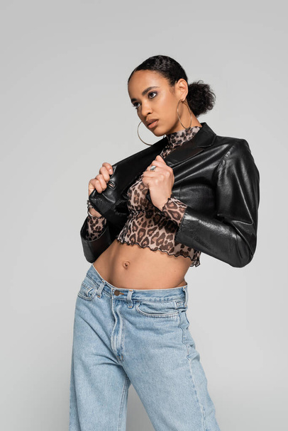 fashionable african american woman adjusting leather cropped jacket while looking away isolated on grey - Fotografie, Obrázek