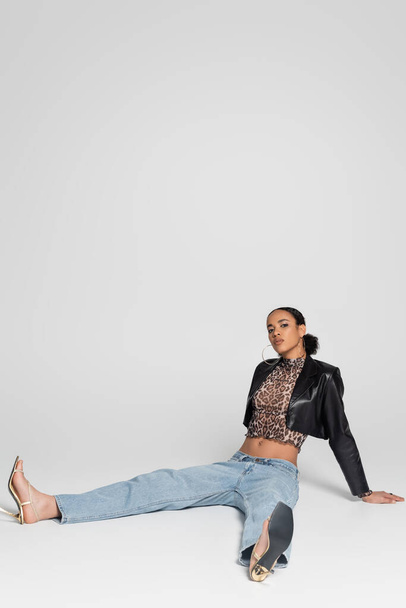 piena lunghezza della giovane donna afroamericana in sandali con tacco alto e vestito alla moda seduta sul grigio  - Foto, immagini