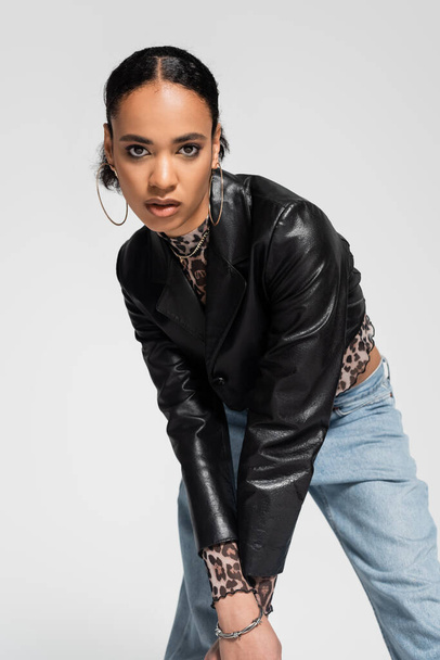fashionable african american woman in trendy cropped jacket and blue jeans looking at camera isolated on grey  - Valokuva, kuva