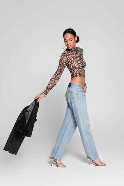 full length of well dressed african american model in crop top with animal print holding black jacket and walking on grey  - Photo, Image