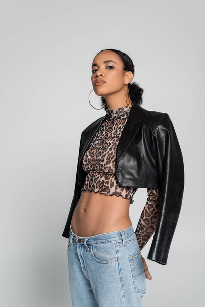 stylish african american model in crop top with animal print and black leather jacket posing isolated on grey  - Foto, imagen