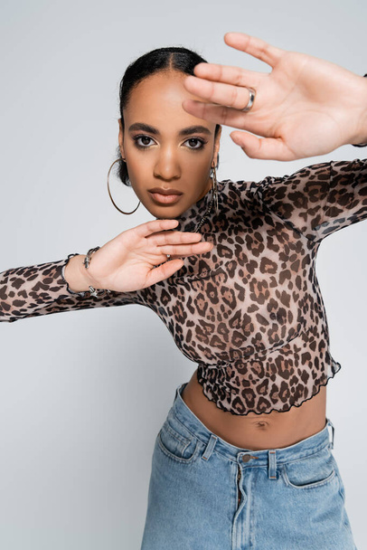 stylish african american woman in crop top with animal print and denim jeans gesturing isolated on grey  - Foto, Bild