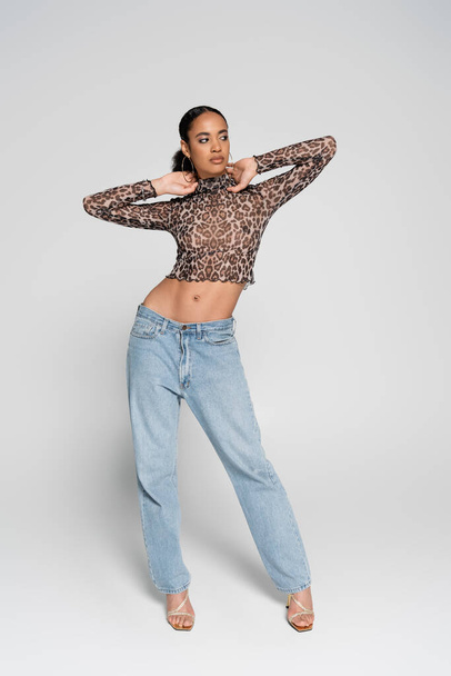 full length of brunette african american woman in crop top with animal print and denim jeans posing on grey  - Zdjęcie, obraz