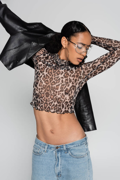 stylish african american woman in crop top with animal print and trendy sunglasses wearing leather jacket isolated on grey  - Fotografie, Obrázek