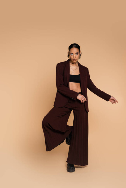 full length of young african american woman in stylish suit with wide pants standing on one leg while posing on beige  - Foto, Imagem