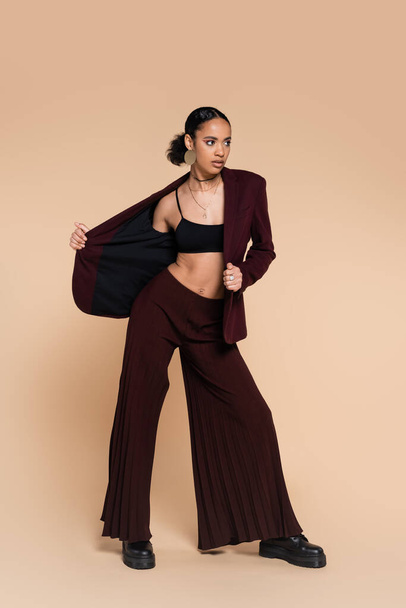 full length of young african american woman in maroon suit taking off stylish blazer on beige  - Photo, Image
