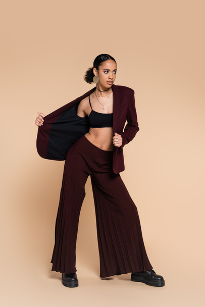 full length of young african american woman in maroon suit taking off stylish blazer on beige  - Fotoğraf, Görsel