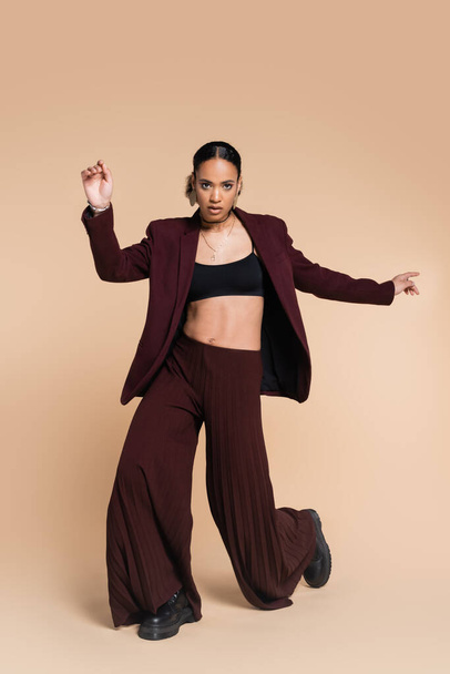 full length of young african american model in maroon suit with wide leg pants posing on beige  - Фото, изображение