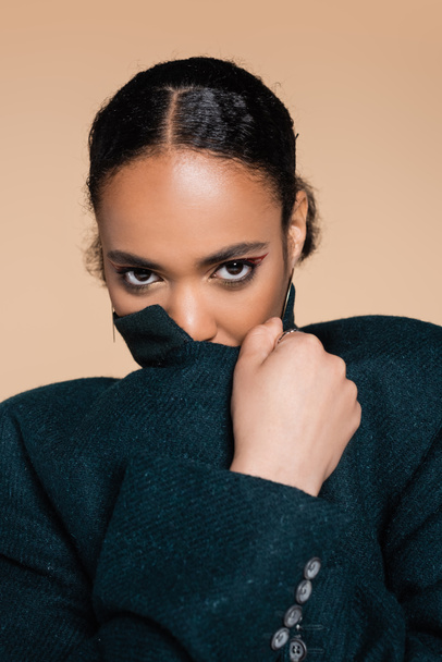 portrait of young african american woman covering face with collar of oversized blazer isolated on beige  - Photo, Image