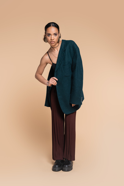 full length of stylish african american model in oversized blazer on shoulder posing with hand on hip on beige  - Photo, Image