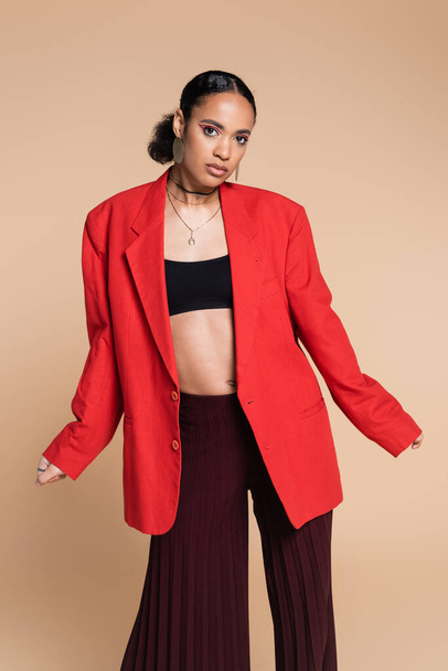 stylish african american model in crop top and red blazer posing isolated on beige  - Photo, Image