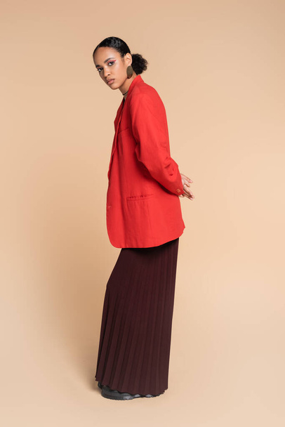 full length of stylish african american model in wide leg pants and red blazer posing on beige  - 写真・画像