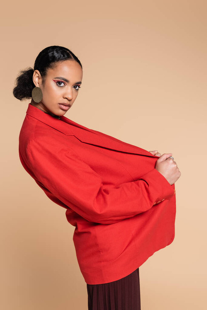 stylish african american model adjusting red blazer while posing isolated on beige  - Photo, Image