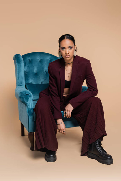 stylish african american model in maroon suit sitting on blue velvet armchair on beige  - Photo, Image