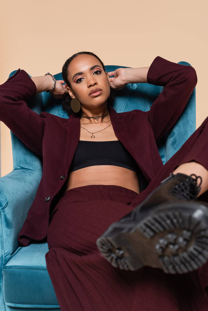 young african american model in stylish suit showing black boot while sitting on blue velvet armchair isolated on beige  - Photo, Image