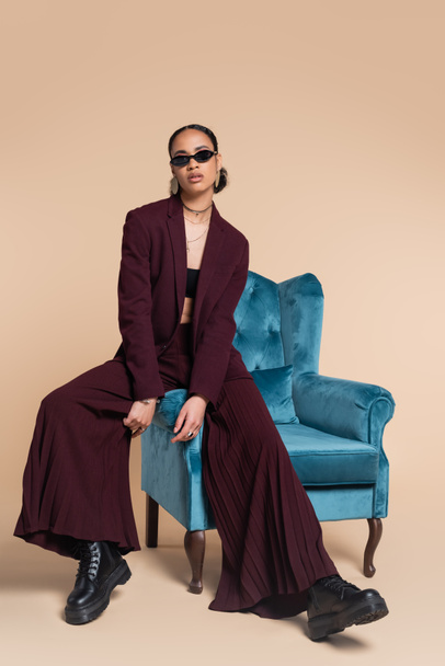 full length of stylish african american woman in burgundy suit and trendy sunglasses sitting on blue velvet armchair on beige  - Valokuva, kuva