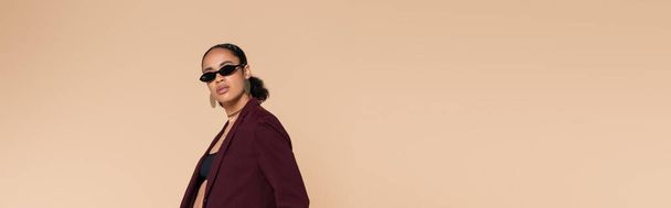 stylish african american woman in burgundy blazer and sunglasses looking at camera isolated on beige, banner  - Foto, afbeelding
