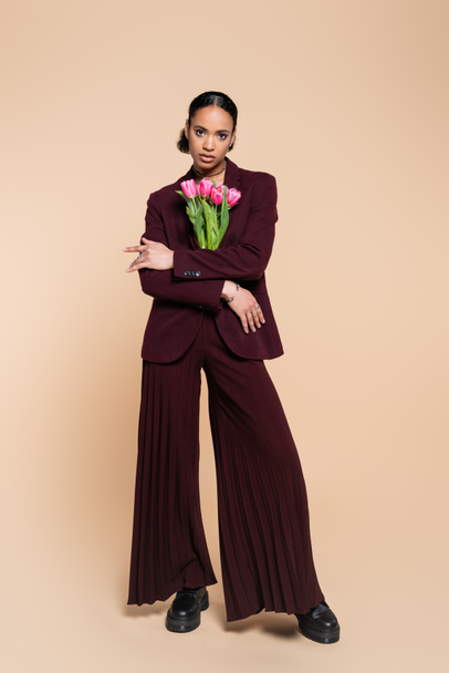 full length of well dressed african american woman in burgundy suit posing with pink tulips on beige  - Фото, изображение