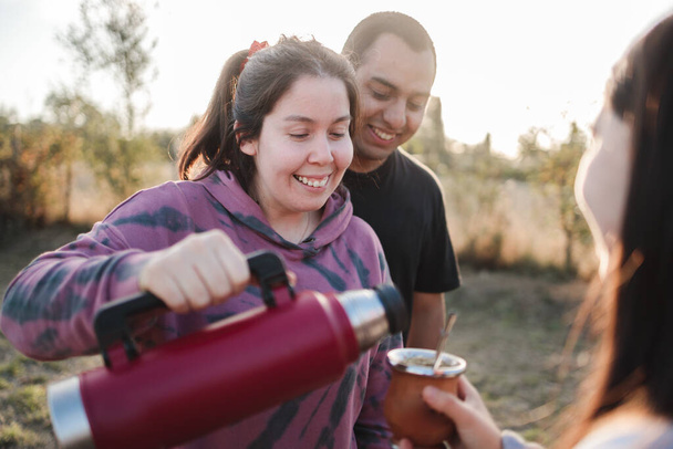 Dos amigos sonrientes bebiendo yerba mate usando un termo con agua caliente en el campo al atardecer. Foto de alta calidad - Foto, Imagen